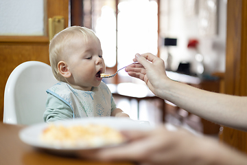 Image showing Mother spoon feeding her infant baby boy child sitting in high chair at the dining table in kitchen at home