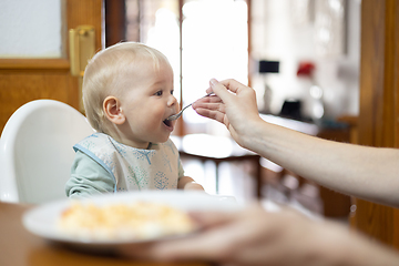 Image showing Mother spoon feeding her infant baby boy child sitting in high chair at the dining table in kitchen at home