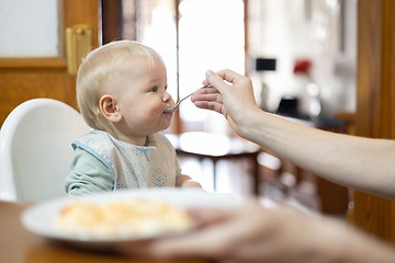 Image showing Mother spoon feeding her infant baby boy child sitting in high chair at the dining table in kitchen at home