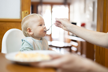 Image showing Mother spoon feeding her infant baby boy child sitting in high chair at the dining table in kitchen at home