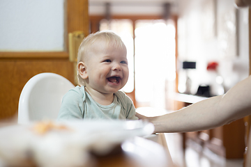 Image showing Adorable cheerful happy infant baby boy child smiling while sitting in high chair at the dining table in kitchen at home beeing spoon fed by his mother