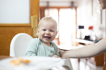 Image showing Adorable cheerful happy infant baby boy child smiling while sitting in high chair at the dining table in kitchen at home beeing spoon fed by his mother.