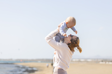 Image showing Mother enjoying summer vacations holding, playing and lifting his infant baby boy son high in the air on sandy beach on Lanzarote island, Spain. Family travel and vacations concept.