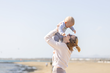 Image showing Mother enjoying summer vacations holding, playing and lifting his infant baby boy son high in the air on sandy beach on Lanzarote island, Spain. Family travel and vacations concept.