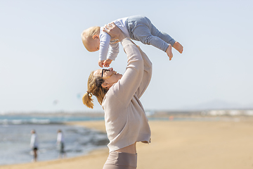 Image showing Mother enjoying summer vacations holding, playing and lifting his infant baby boy son high in the air on sandy beach on Lanzarote island, Spain. Family travel and vacations concept.