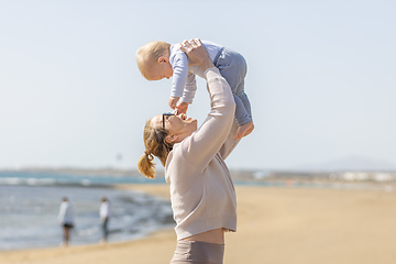 Image showing Mother enjoying summer vacations holding, playing and lifting his infant baby boy son high in the air on sandy beach on Lanzarote island, Spain. Family travel and vacations concept.