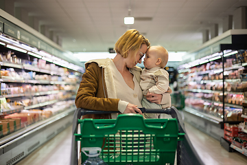 Image showing Mother pushing shopping cart with her infant baby boy child down department aisle in supermarket grocery store. Shopping with kids concept.