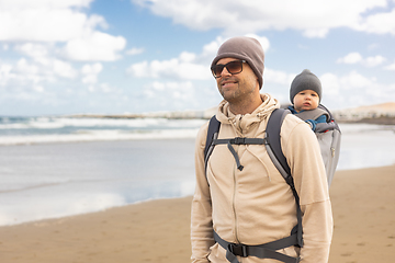 Image showing Young father carrying his infant baby boy son in backpack on windy sandy beach. Family travel and winter vacation concept.