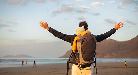 Image showing Young father rising hands to the sky while enjoying pure nature carrying his infant baby boy son in backpack on windy sandy beach of Famara, Lanzarote island, Spain at sunset. Family travel concept.