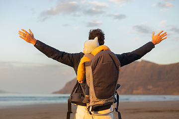 Image showing Young father rising hands to the sky while enjoying pure nature carrying his infant baby boy son in backpack on windy sandy beach of Famara, Lanzarote island, Spain at sunset. Family travel concept.