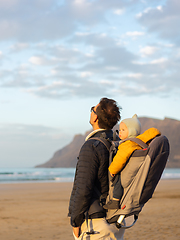 Image showing Young father rising hands to the sky while enjoying pure nature carrying his infant baby boy son in backpack on windy sandy beach of Famara, Lanzarote island, Spain at sunset. Family travel concept.