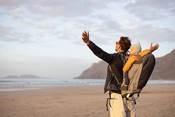 Image showing Young father rising hands to the sky while enjoying pure nature carrying his infant baby boy son in backpack on windy sandy beach of Famara, Lanzarote island, Spain at sunset. Family travel concept.