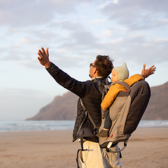 Image showing Young father rising hands to the sky while enjoying pure nature carrying his infant baby boy son in backpack on windy sandy beach of Famara, Lanzarote island, Spain at sunset. Family travel concept.