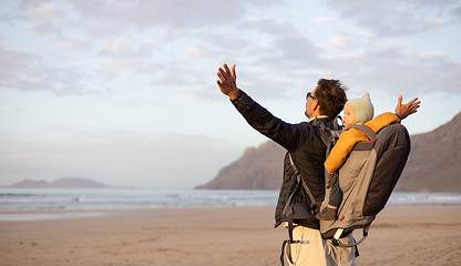 Image showing Young father rising hands to the sky while enjoying pure nature carrying his infant baby boy son in backpack on windy sandy beach of Famara, Lanzarote island, Spain at sunset. Family travel concept.