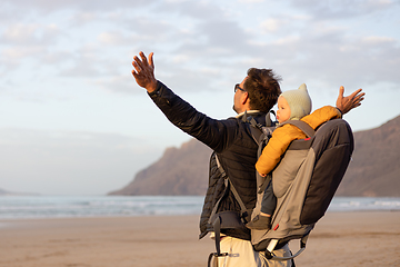 Image showing Young father rising hands to the sky while enjoying pure nature carrying his infant baby boy son in backpack on windy sandy beach of Famara, Lanzarote island, Spain at sunset. Family travel concept.