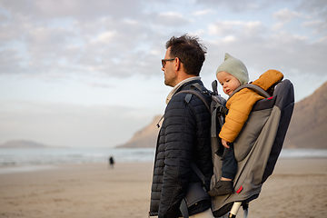 Image showing Father enjoying pure nature carrying his infant baby boy son in backpack on windy sandy beach of Famara, Lanzarote island, Spain at sunset. Family travel concept.