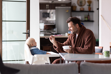 Image showing Father wearing bathrope spoon feeding hir infant baby boy child sitting in high chair at the dining table in kitchen at home in the morning.