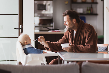 Image showing Father wearing bathrope spoon feeding hir infant baby boy child sitting in high chair at the dining table in kitchen at home in the morning.