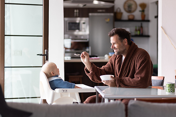 Image showing Father wearing bathrope spoon feeding hir infant baby boy child sitting in high chair at the dining table in kitchen at home in the morning.