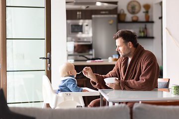 Image showing Father wearing bathrope spoon feeding hir infant baby boy child sitting in high chair at the dining table in kitchen at home in the morning.