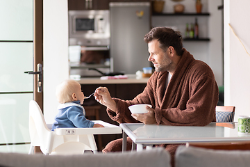 Image showing Father wearing bathrope spoon feeding hir infant baby boy child sitting in high chair at the dining table in kitchen at home in the morning.