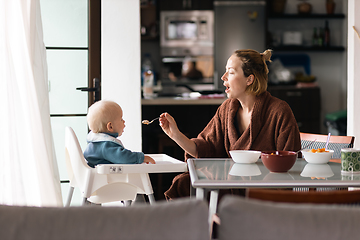 Image showing Cheerful mother wearing bathrope spoon feeding her infant baby boy child sitting in high chair at the dining table in kitchen at home in the morning.