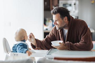 Image showing Father wearing bathrope spoon feeding hir infant baby boy child sitting in high chair at the dining table in kitchen at home in the morning.