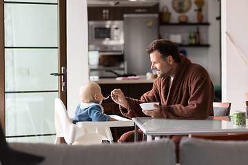 Image showing Father wearing bathrope spoon feeding hir infant baby boy child sitting in high chair at the dining table in kitchen at home in the morning.