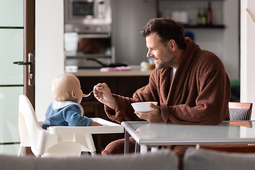 Image showing Father wearing bathrope spoon feeding hir infant baby boy child sitting in high chair at the dining table in kitchen at home in the morning.