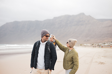 Image showing Young father carrying his infant baby boy son in backpack on windy sandy beach. Family travel and winter vacation concept.