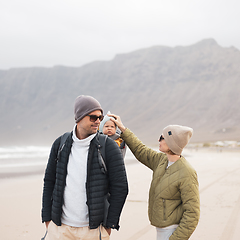 Image showing Young father carrying his infant baby boy son in backpack on windy sandy beach. Family travel and winter vacation concept.