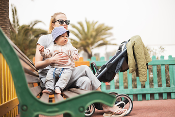 Image showing Young mother with her cute infant baby boy child on bench on urban children's playground on warm summer day.