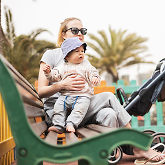 Image showing Young mother with her cute infant baby boy child on bench on urban children's playground on warm summer day.