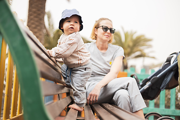 Image showing Young mother with her cute infant baby boy child on bench on urban children's playground on warm summer day.