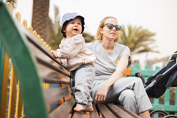 Image showing Young mother with her cute infant baby boy child on bench on urban children's playground on warm summer day.