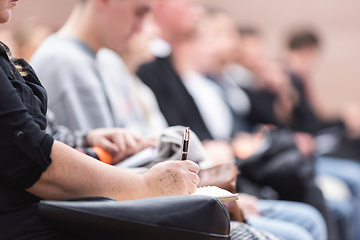 Image showing Female hands holding pen and notebook, making notes at conference lecture. Event participants in conference hall.