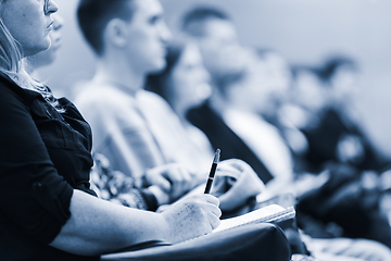 Image showing Female hands holding pen and notebook, making notes at conference lecture. Event participants in conference hall. Blue toned greyscale image.