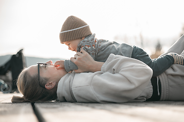 Image showing Happy family. Young mother playing with her baby boy infant oudoors on sunny autumn day. Portrait of mom and little son on wooden platform by lake. Positive human emotions, feelings, joy.