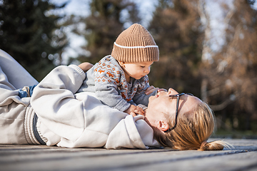 Image showing Happy family. Young mother playing with her baby boy infant oudoors on sunny autumn day. Portrait of mom and little son on wooden platform by lake. Positive human emotions, feelings, joy.