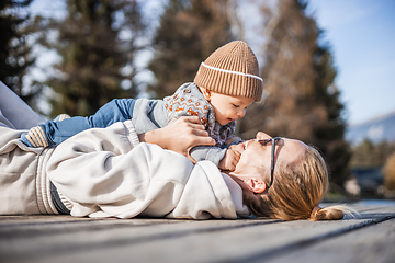 Image showing Happy family. Young mother playing with her baby boy infant oudoors on sunny autumn day. Portrait of mom and little son on wooden platform by lake. Positive human emotions, feelings, joy.