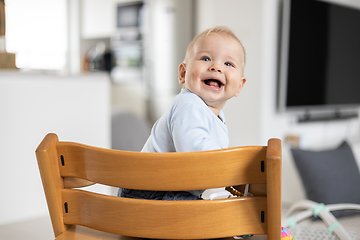 Image showing Happy infant sitting in traditional scandinavian designer wooden high chair and laughing out loud in modern bright home. Cute baby smile.