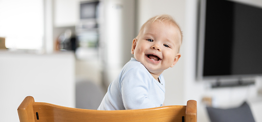 Image showing Happy infant sitting in traditional scandinavian designer wooden high chair and laughing out loud in modern bright home. Cute baby smile.