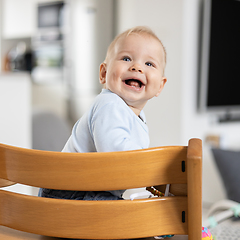 Image showing Happy infant sitting in traditional scandinavian designer wooden high chair and laughing out loud in modern bright home. Cute baby smile.