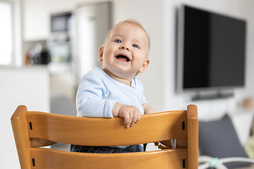 Image showing Happy infant sitting in traditional scandinavian designer wooden high chair and laughing out loud in modern bright home. Cute baby smile.