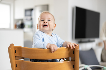 Image showing Happy infant sitting in traditional scandinavian designer wooden high chair and laughing out loud in modern bright home. Cute baby smile.