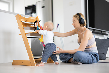 Image showing Women's multitasking. Mother sitting on floor playing with her baby boy watching and suppervising his first steps while listening to podcast on wireless headphones.