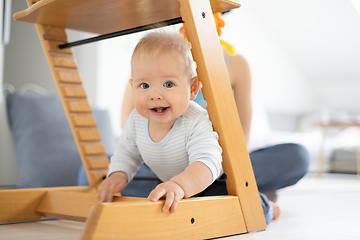 Image showing Happy infant climbing under traditional scandinavian designer wooden high chair and in modern bright home. Cute baby smiling in camera