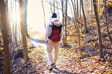 Image showing Sporty father carrying his infant son wearing winter jumpsuit and cap in backpack carrier hiking in autumn forest.