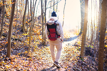 Image showing Sporty father carrying his infant son wearing winter jumpsuit and cap in backpack carrier hiking in autumn forest.