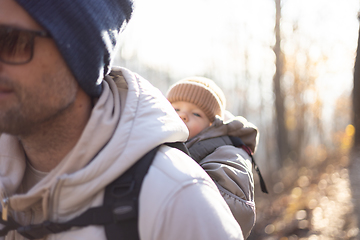 Image showing Sporty father carrying his infant son wearing winter jumpsuit and cap in backpack carrier hiking in autumn forest.